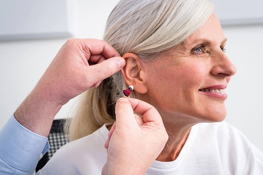 Seniorin bekommt ein modernes Hörgerät angepasst, Hörakustiker unterstützt beim Einsetzen.